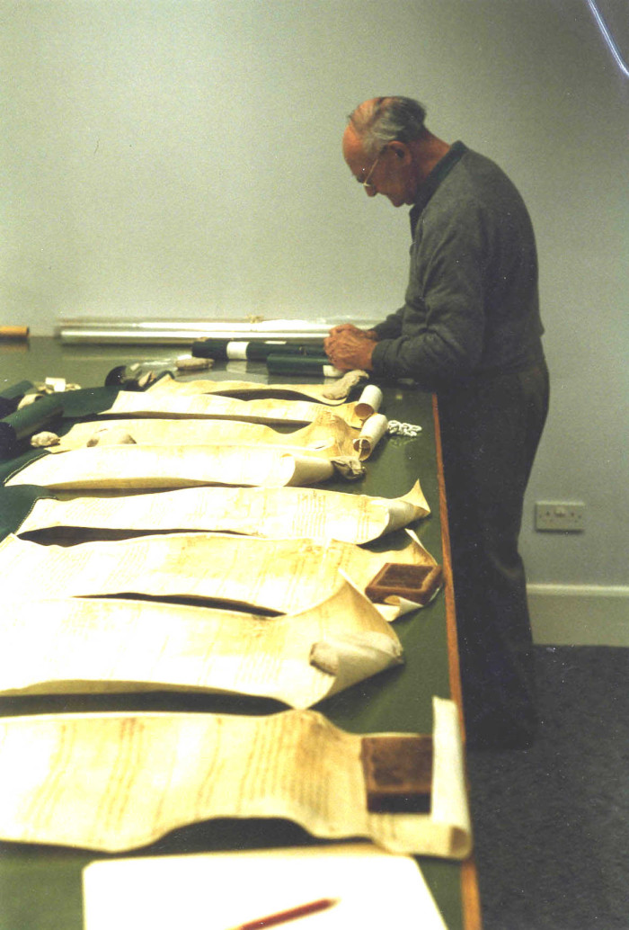 A man stands at a bench table with parchment documents along it
