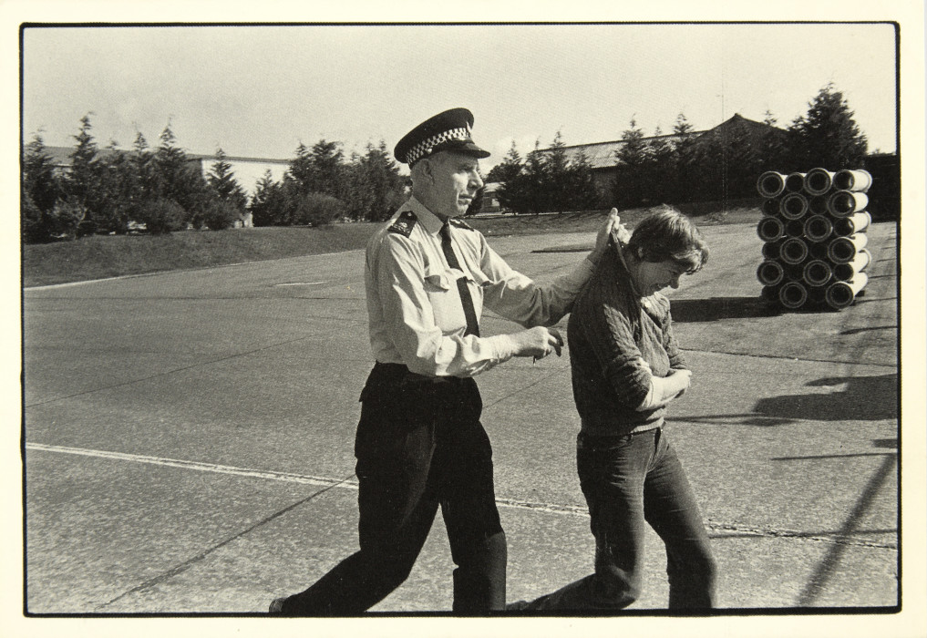 Postcard of a man (possibly police or security guard) holding a person (a woman?) by the scruff of their clothing, 1983. From Greenham Common POSTCARDS/ collection.